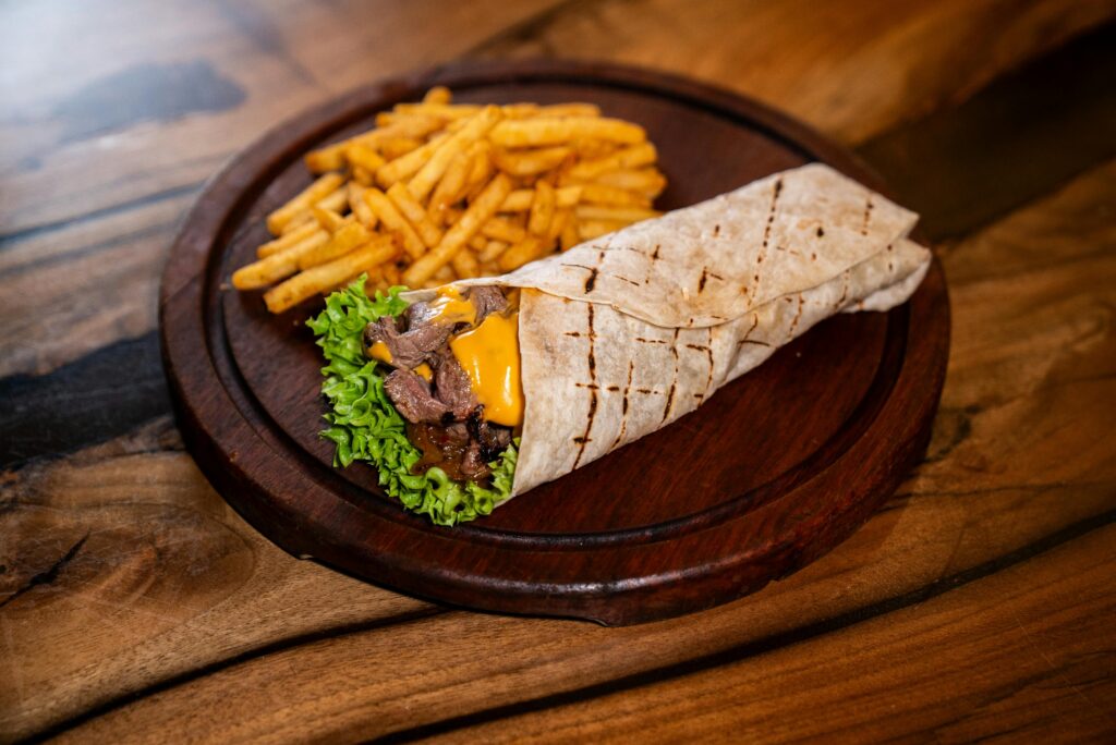 Delicious beef wrap with cheddar, lettuce and fries on wooden plate. Perfect for fast food lovers.